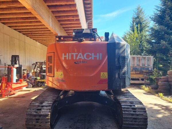 A large orange Hitachi excavator parked inside a wooden garage with various tools and equipment around