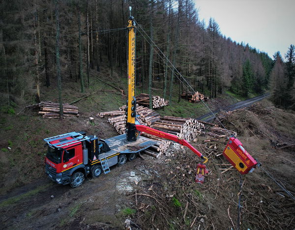Aerial view of the Mounty 5000 crane mounted on a truck, lifting logs in a forest with multiple stacks of wood in the background.